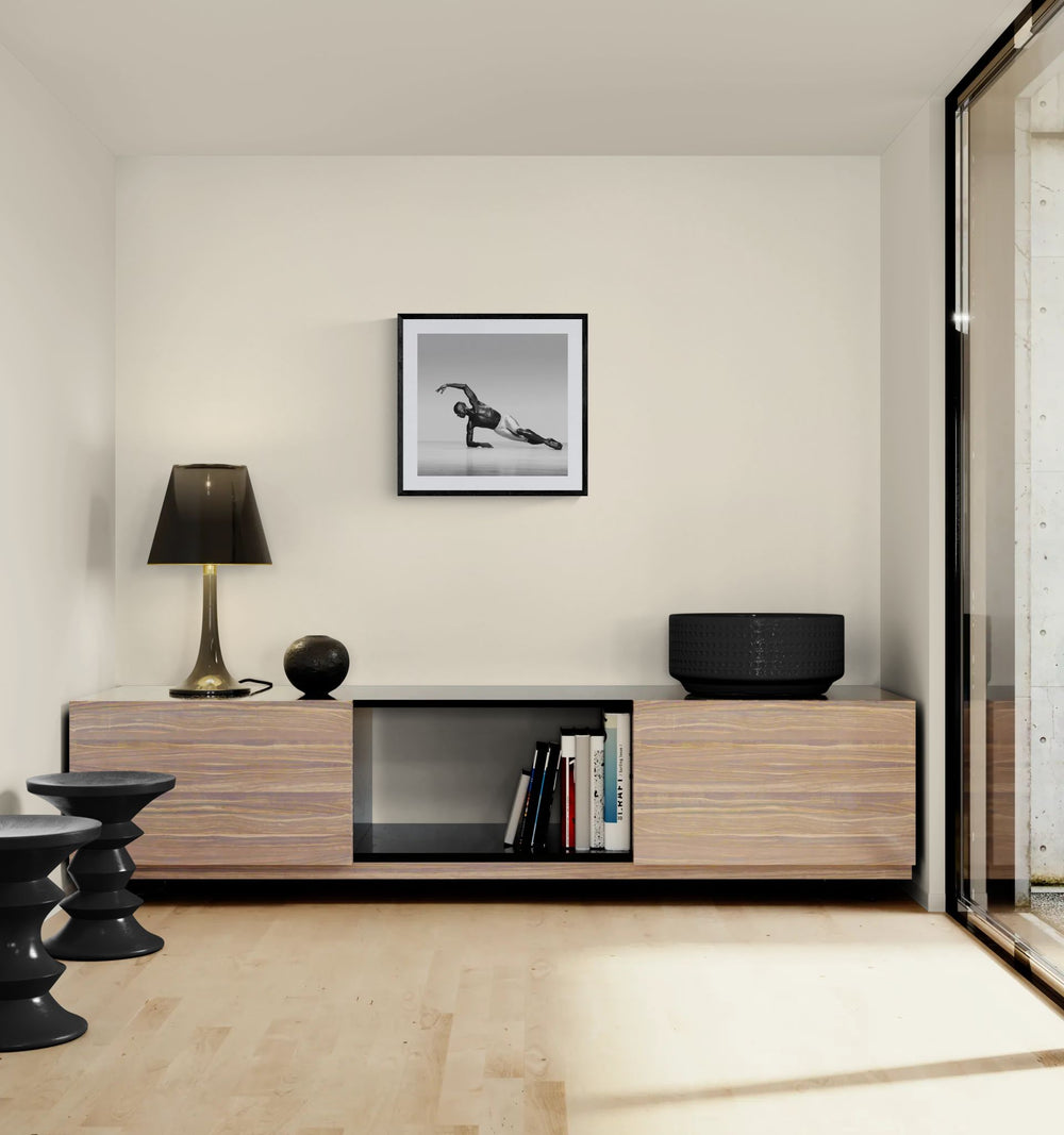 Modern living room corner with a low wooden console, lamp and decor objects, featuring a framed square black-and-white dance photograph on the wall above.