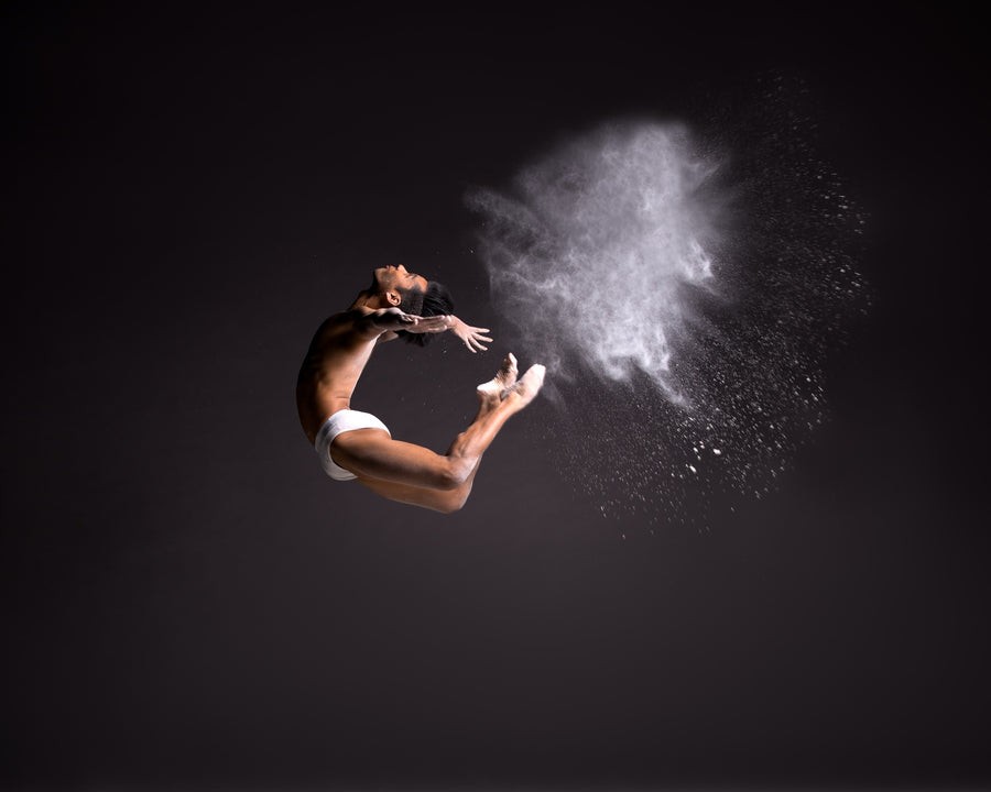 Male dancer in mid-air leap against a dark background, arched backwards with arms and legs extended as a white powder cloud bursts in front of him.