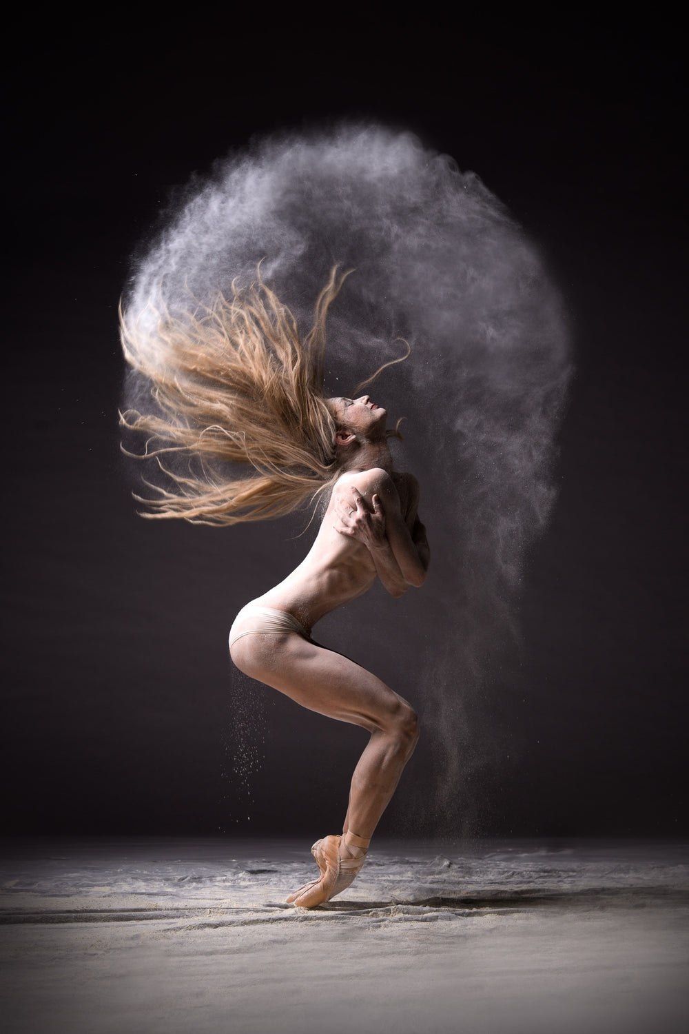 Female dancer on pointe folded inward, hair and white powder forming a halo arc above her head on a dark stage.