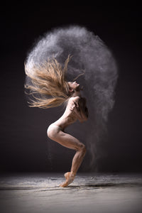 Female dancer on pointe folded inward, hair and white powder forming a halo arc above her head on a dark stage.