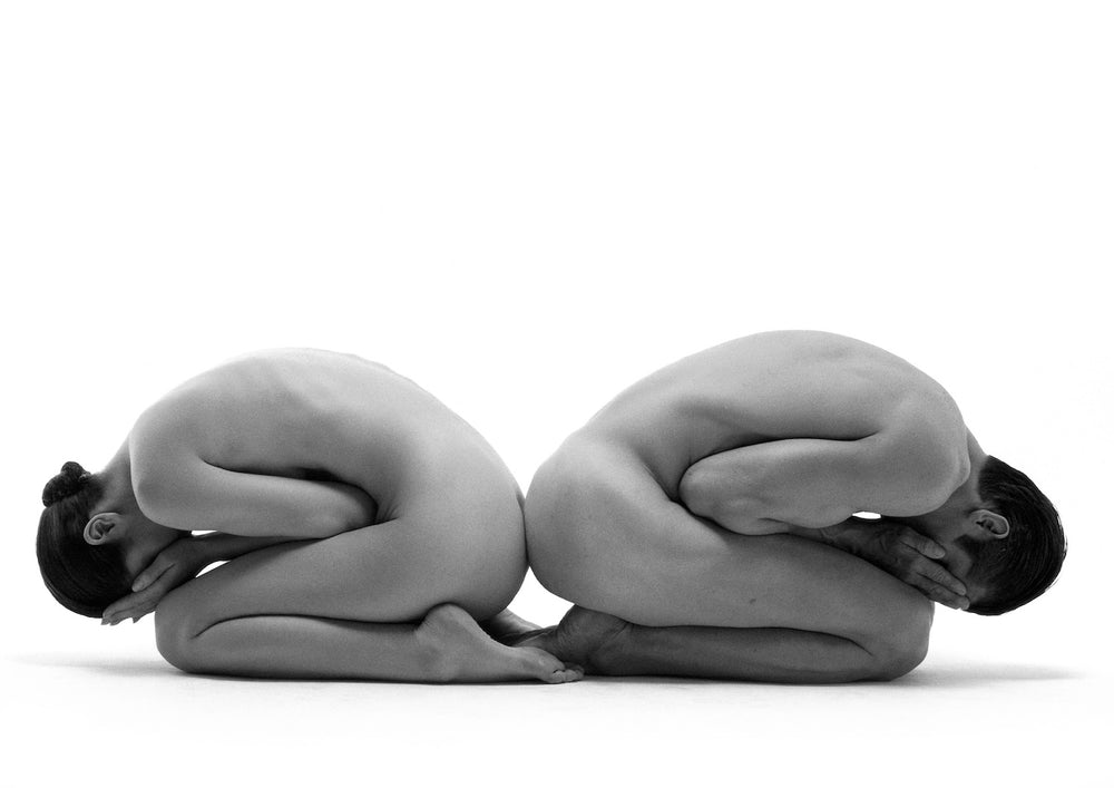 Two nude dancers kneeling back to back on a white field, bodies folded into mirrored curves that form one sculptural shape.