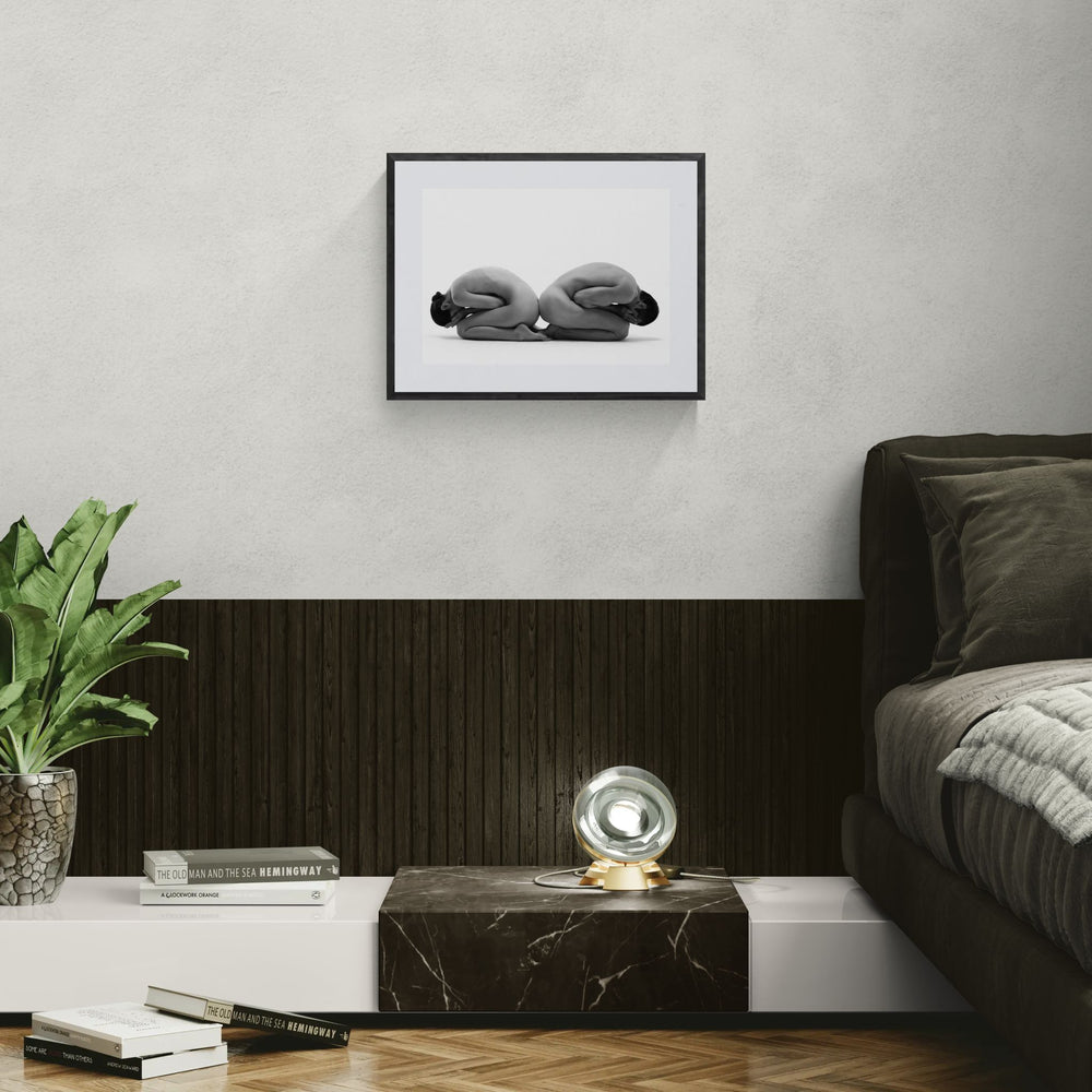 Framed Whitebound Resonance nude dance print hanging above a low bedside platform with dark wood panelling and a green plant in a modern bedroom.