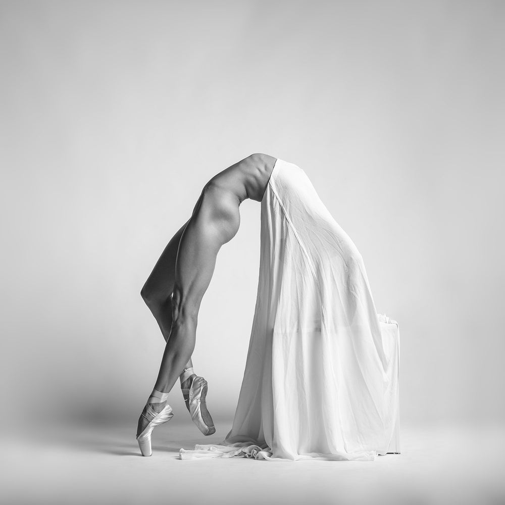 A powerful and graceful black-and-white ballet fine art photograph by Arkadiusz Branicki, featuring a ballerina in a deep arch on pointe, elegantly draped with white fabric. The dancer's muscular form contrasts beautifully with the softness of the fabric, highlighting strength and grace.