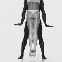 Black-and-white photograph of two dancers aligned one in front of the other, standing on pointe with arms extended, creating a mirrored vertical figure.