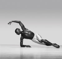 Black-and-white photograph of a male dancer balancing on one forearm with his body extended in a long diagonal line close to the floor, other arm arcing overhead.
