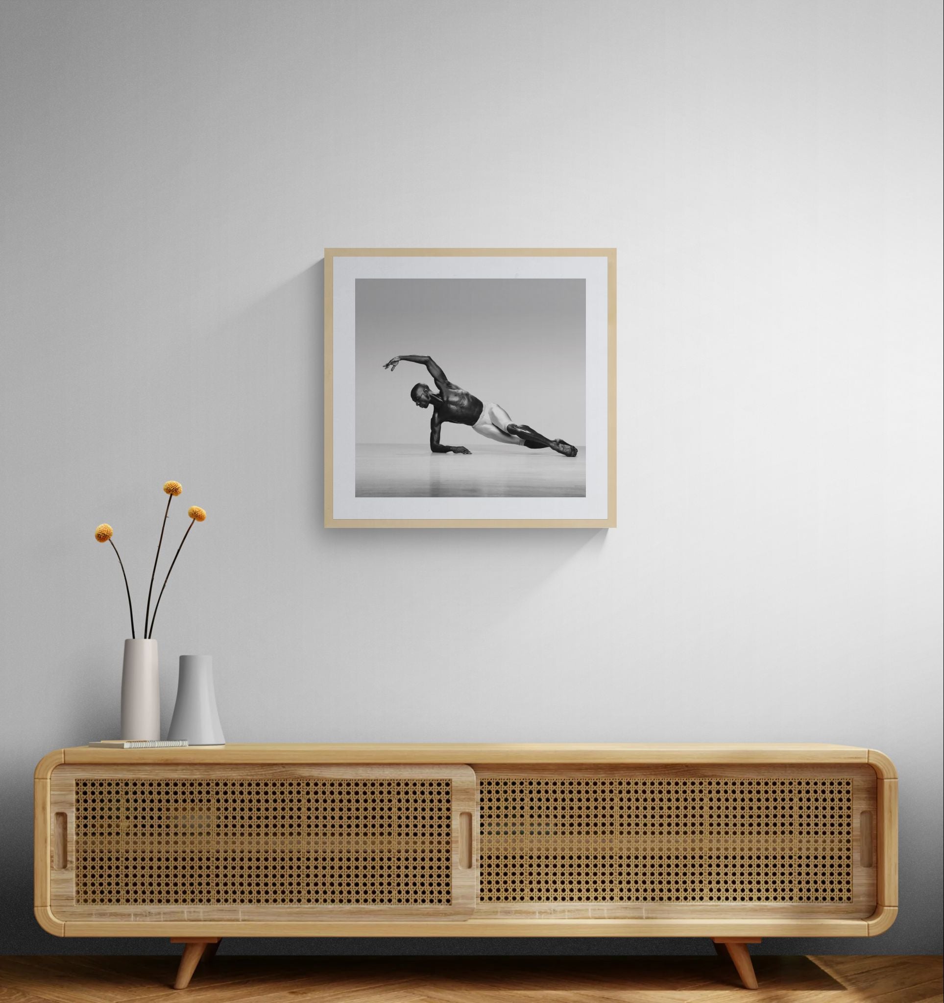 Neutral interior with a cane-front wooden console and slim vases, with a framed square black-and-white dance photograph hanging above.