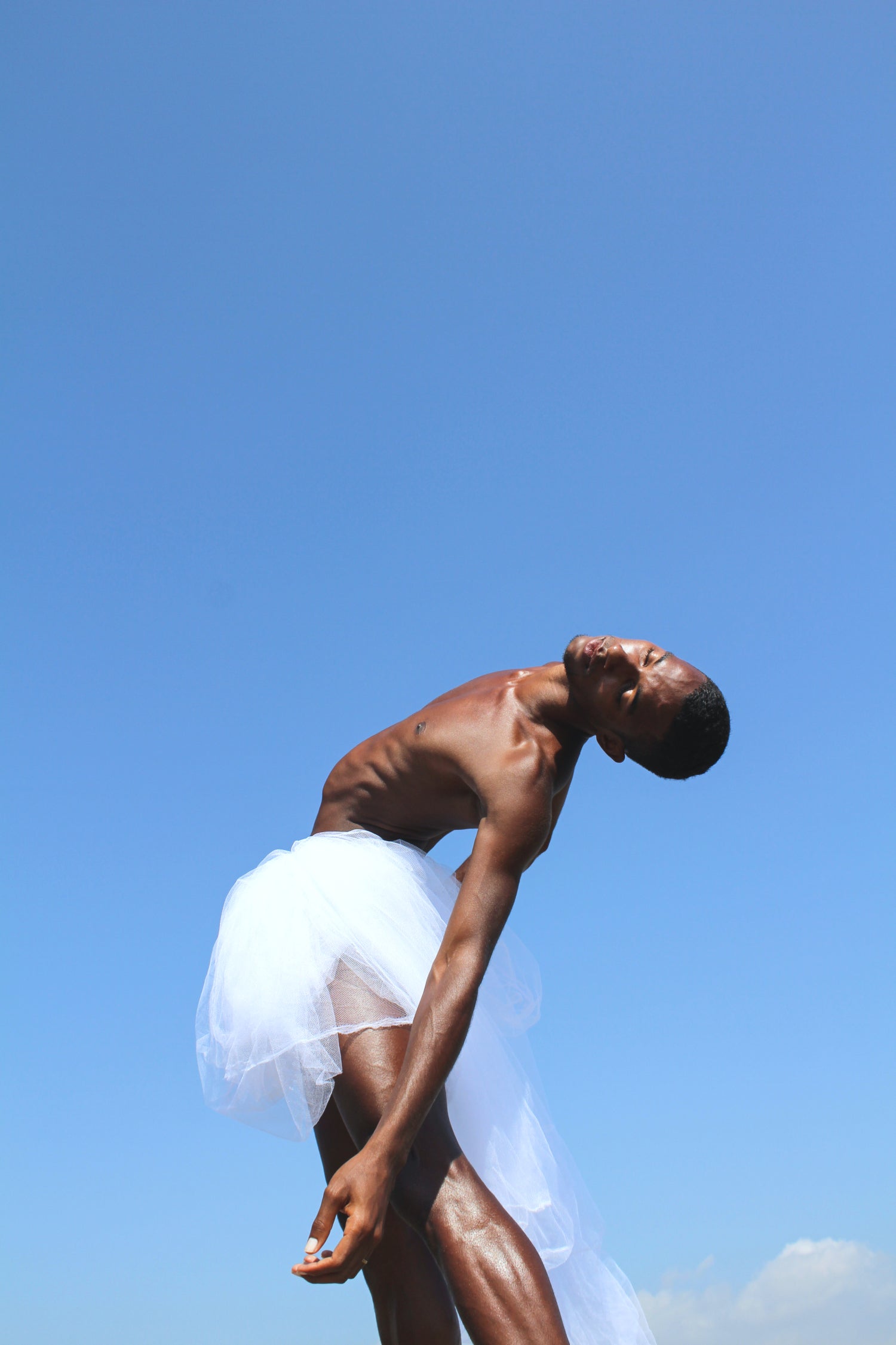 Black male dancer in a meditative movement mood. his skin is glowing into the sun. He is wearing a white skirt on a perfect blue natural sky background.